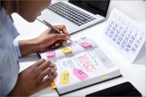 Close-up of a person writing in a planner filled with colorful sticky notes and reminders, next to a desk calendar and an open laptop—symbolizing organized scheduling and checking the Kitimat events calendar.