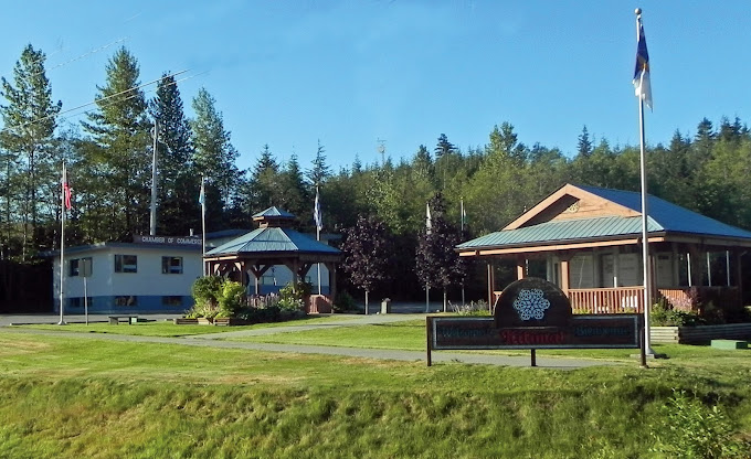View of the Kitimat Chamber of Commerce building and visitor centre on a sunny day, surrounded by flags, gazebos, and greenery, welcoming guests to the community