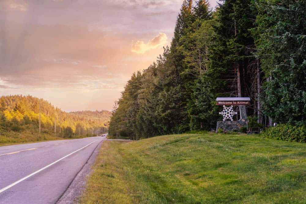 Sunset view of the 'Welcome to Kitimat' roadside sign framed by lush forest, inviting visitors into the community along a scenic northern BC highway.