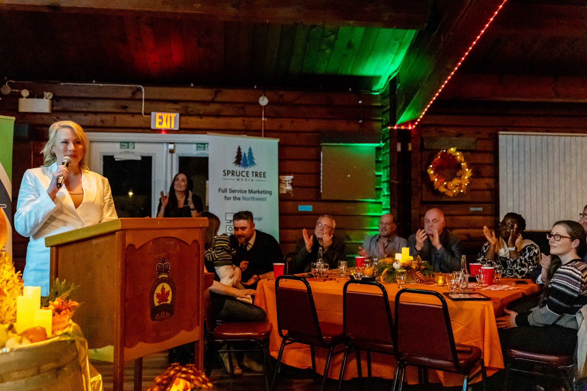 A woman in a white blazer speaks at a podium during the Kitimat Business Excellence Awards, while attendees seated at decorated tables with candles and fall-themed centerpieces applaud in a warmly lit venue