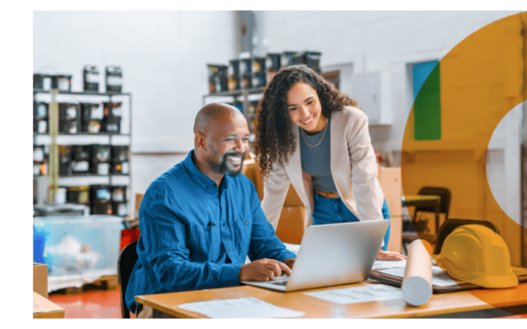 Two small business owners smiling while reviewing plans on a laptop in a warehouse, highlighting collaboration and entrepreneurial support.