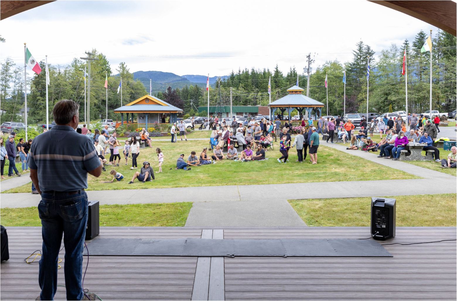 A speaker is addressing a large crowd at an outdoor community event at Heritage Park, Kitimat, with families gathered on the lawn and local flags flying in the background.