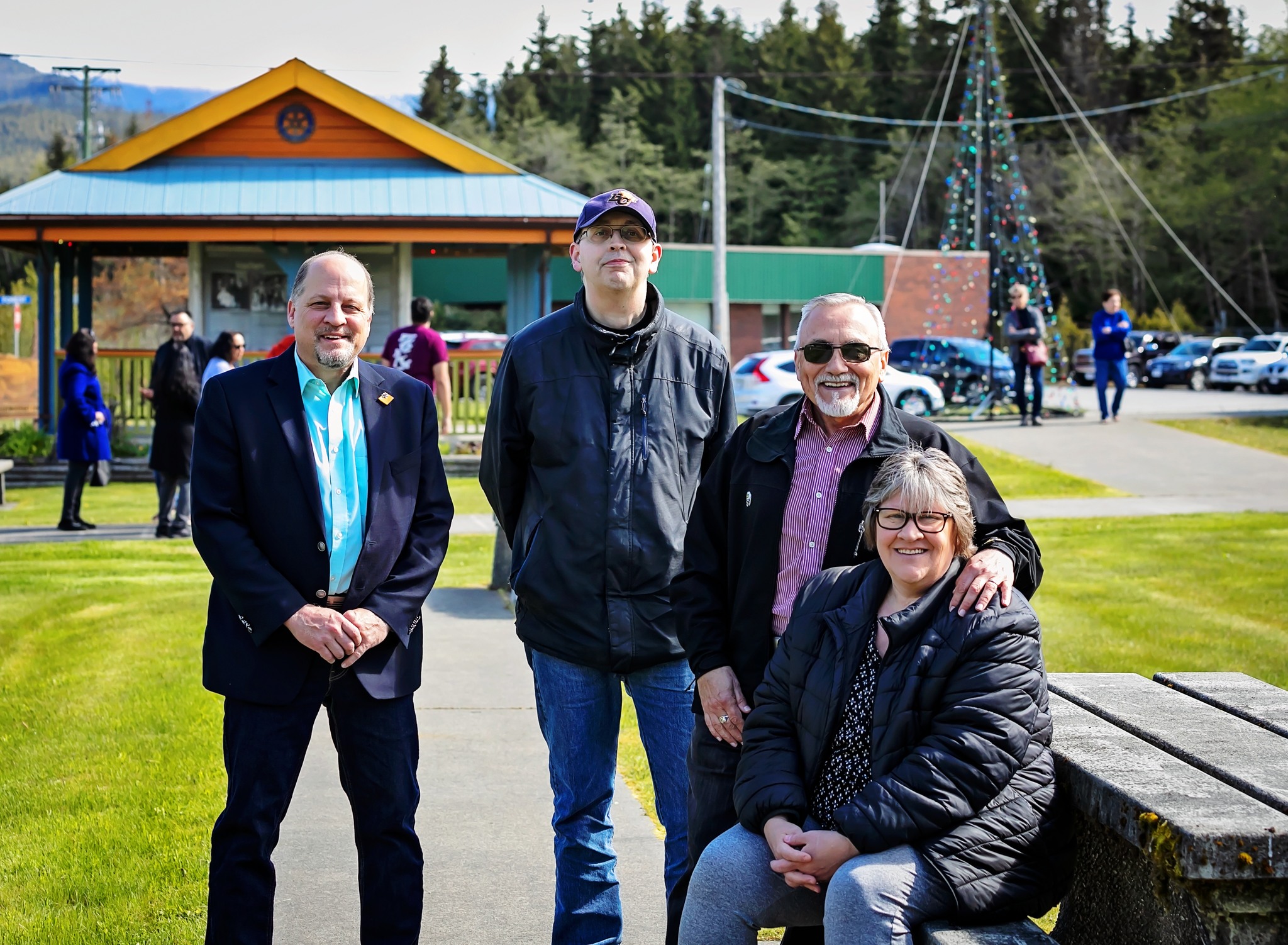 Group of four adults smiling at a community event in a park, with a pavilion and decorated flagpole in the background.