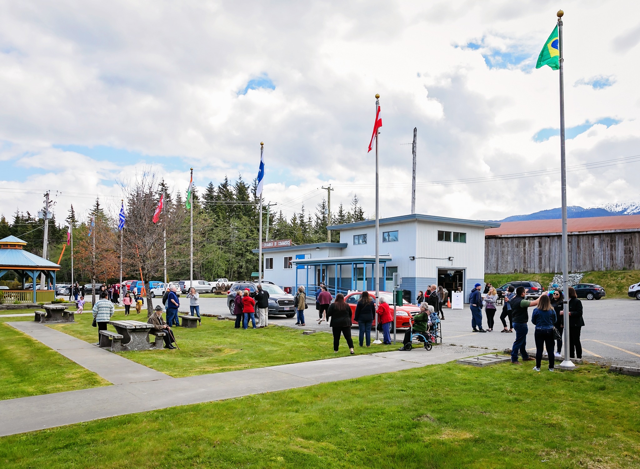 The image shows a gathering of people in a town square, possibly for a community event. Several flags of different nations are visible, suggesting a diverse community or an international event. People are standing in groups, some seated on benches. Cars are parked in the background, and a small building, possibly a community center or Chamber of Commerce, is central to the scene. The overall atmosphere appears peaceful and community-oriented. The presence of children suggests a family-friendly event. The overcast sky adds to the muted, everyday feel of the scene.